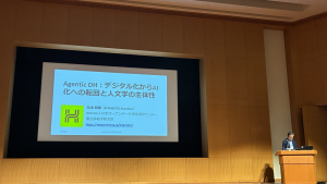 講堂の壇上を撮影した写真。舞台中央のスクリーンに「Agentic DH: デジタル化からAI化への展開と人文学の主体性」という講演タイトルが映し出されていて、右側で北本先生が演壇に立っている。